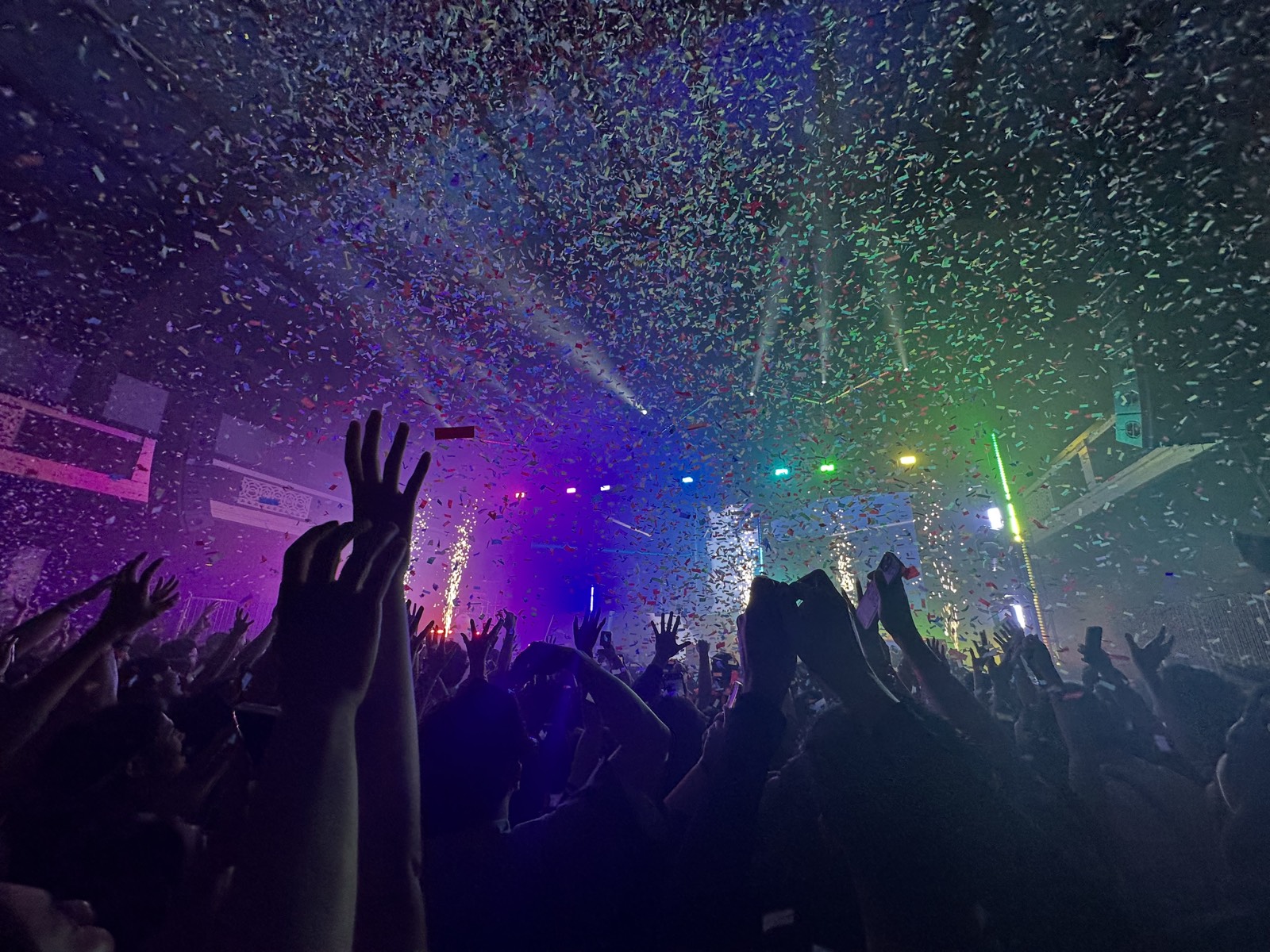 Concert crowd with hands raised under falling confetti and coloured stage lights