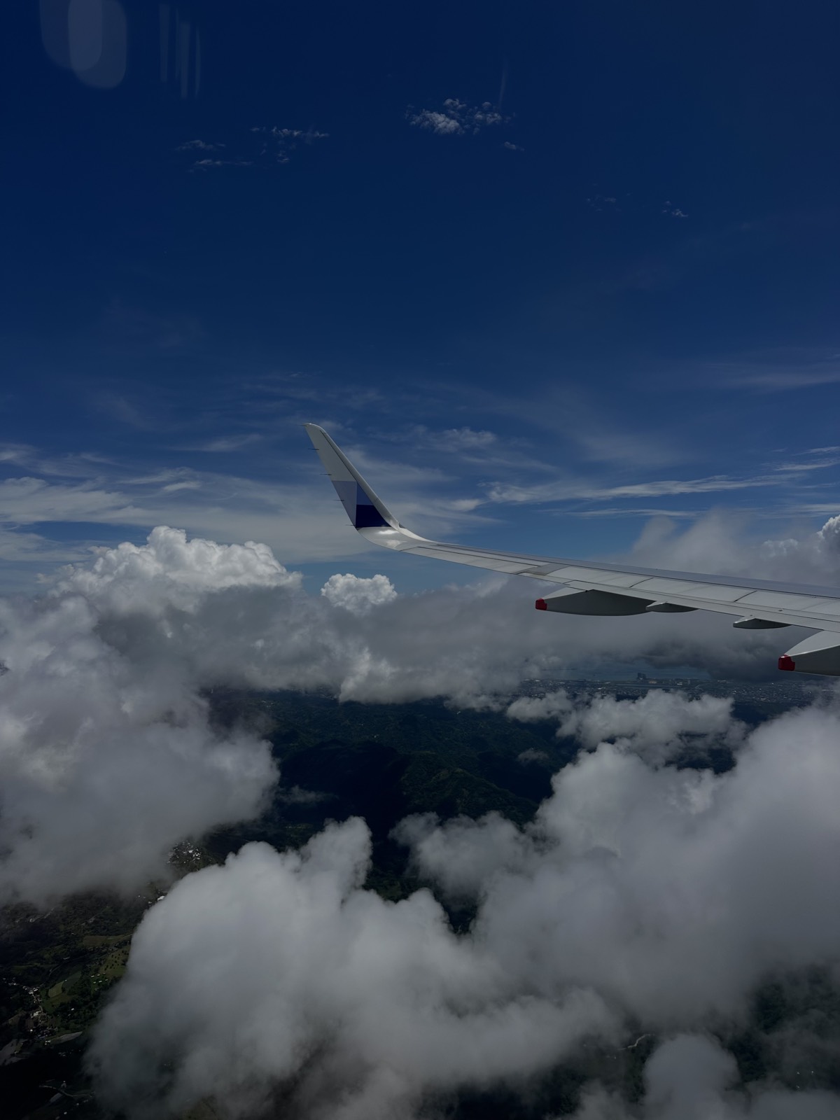 View from an airplane window: the wing above clouds and green hills below